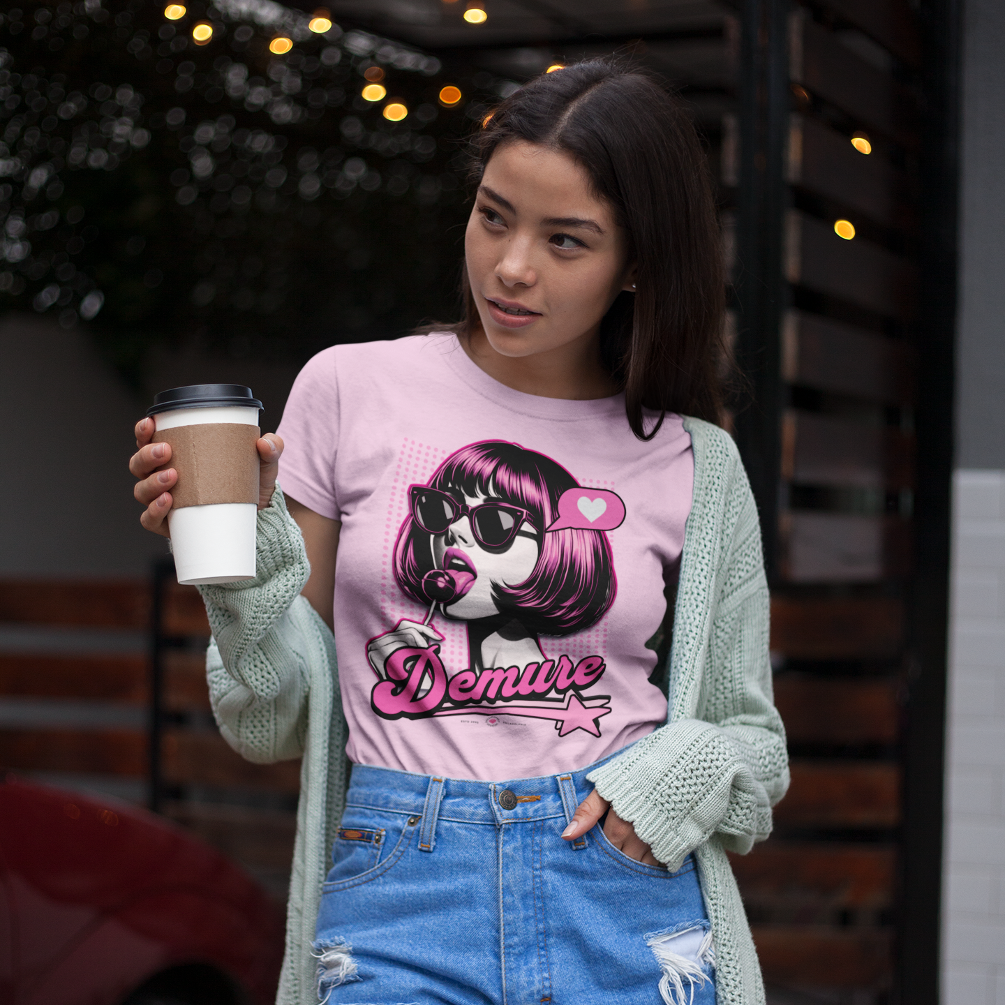 A woman in a pink graphic crop top and a cardigan holding a coffee.